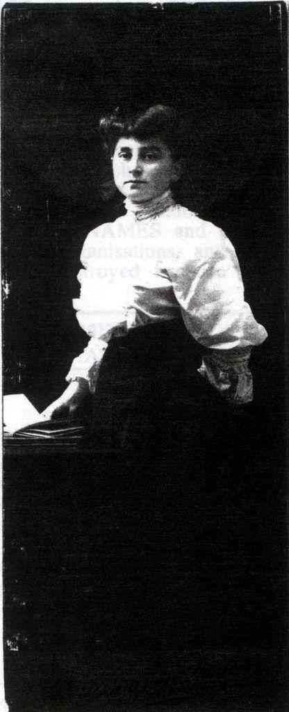 A historical black-and-white photo of a woman standing by a table and posing for the camera in an elegant blouse and necktie, exuding a confident demeanor.
